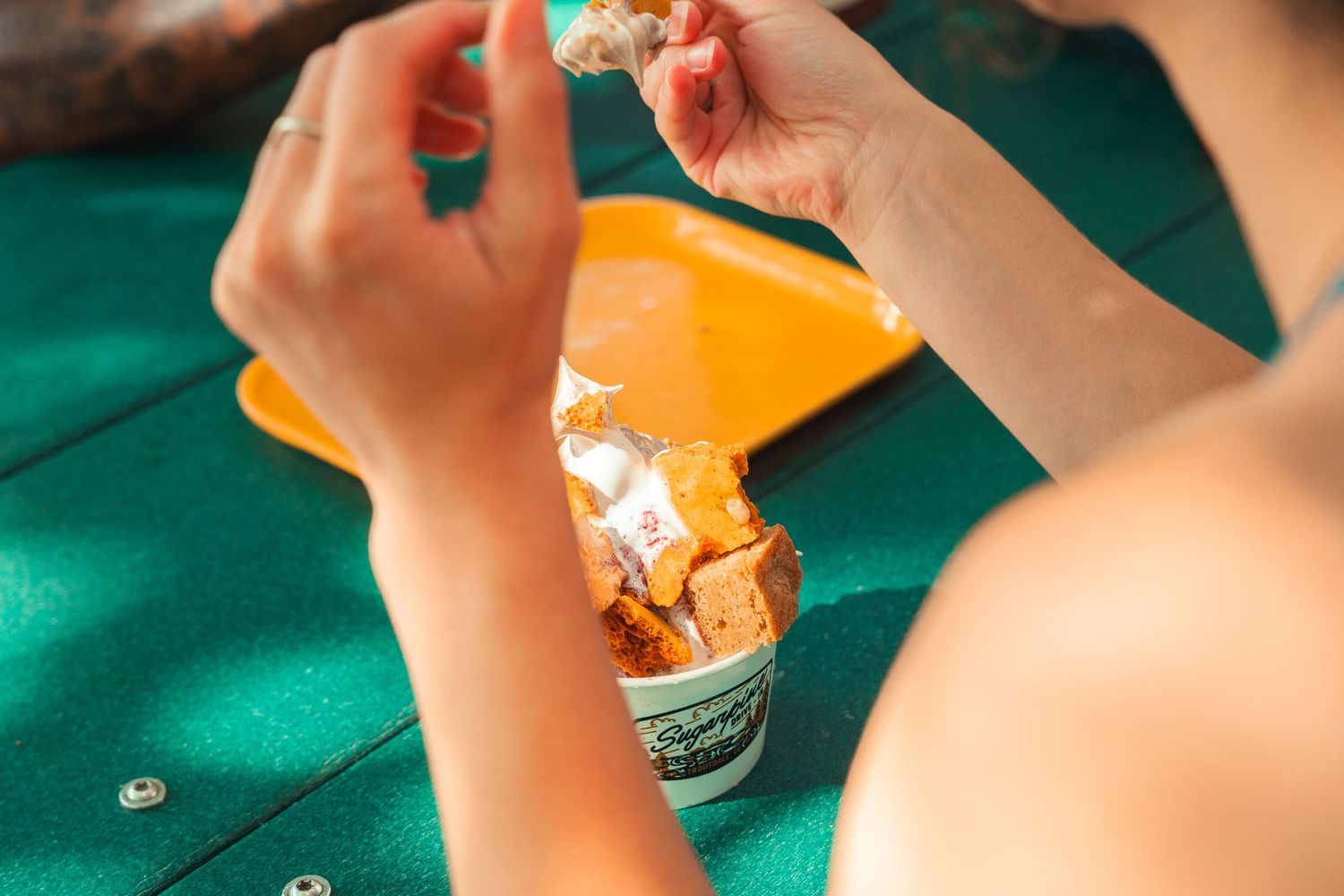 Person eating crispy chicken from a paper cup on a green table outdoors in sunny weather.