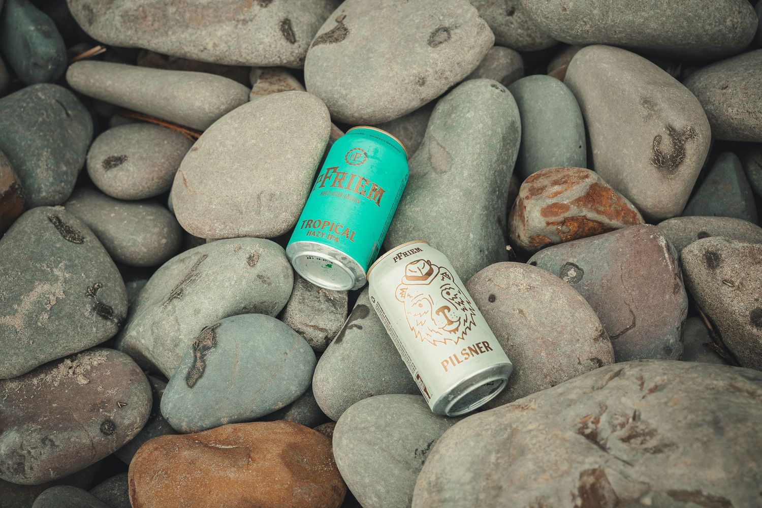 Two beer cans resting on smooth river rocks outdoors, one teal and one white, surrounded by natural grey and brown pebbles.