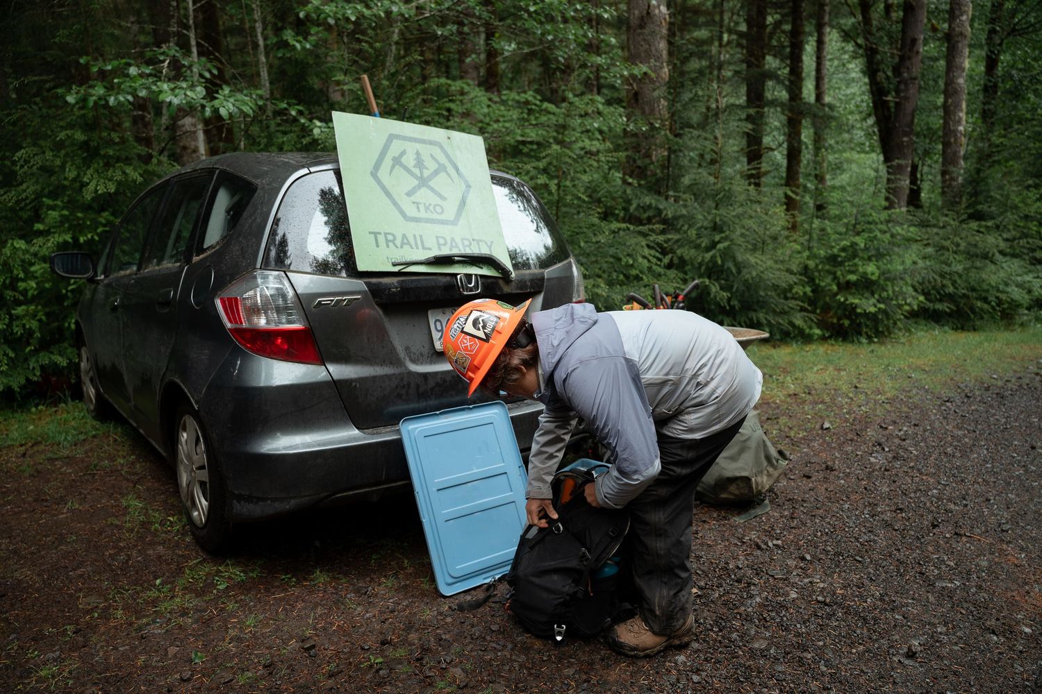 Person crouching behind a Honda Fit hatchback in a forest, with a green trail sign on the car trunk.