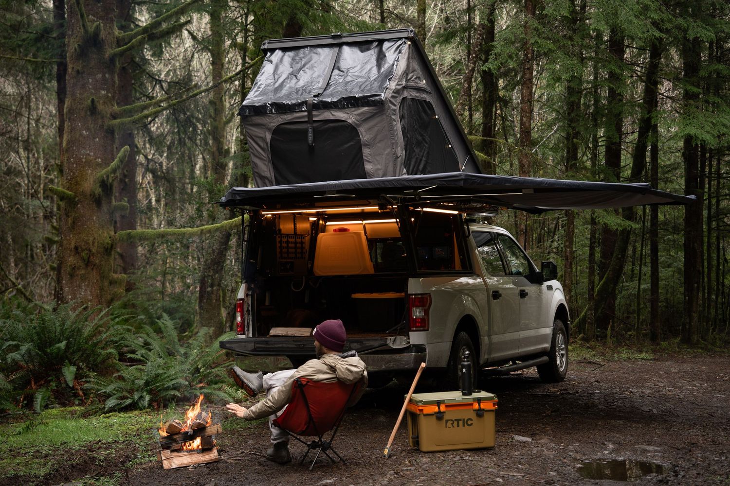 Camping setup with rooftop tent, campfire, and cooler in forest setting at dusk.