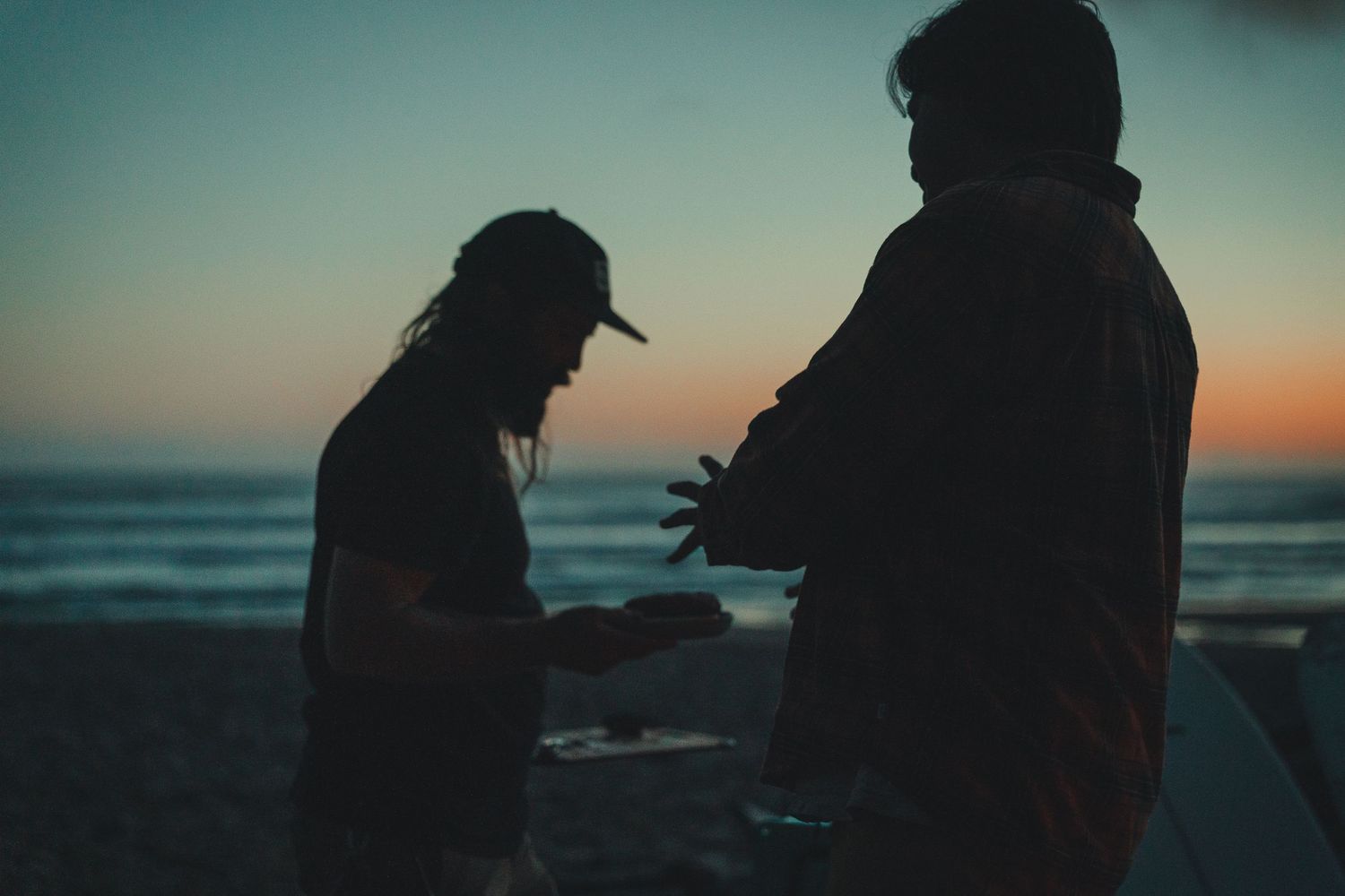 Two silhouetted figures interact on a beach at dusk with a colorful sunset sky over the ocean.