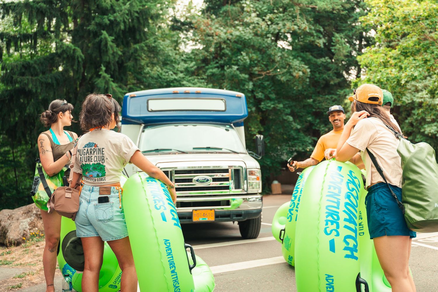Group of friends carrying green inflatable tubes toward a shuttle bus for a river tubing adventure on a sunny day.