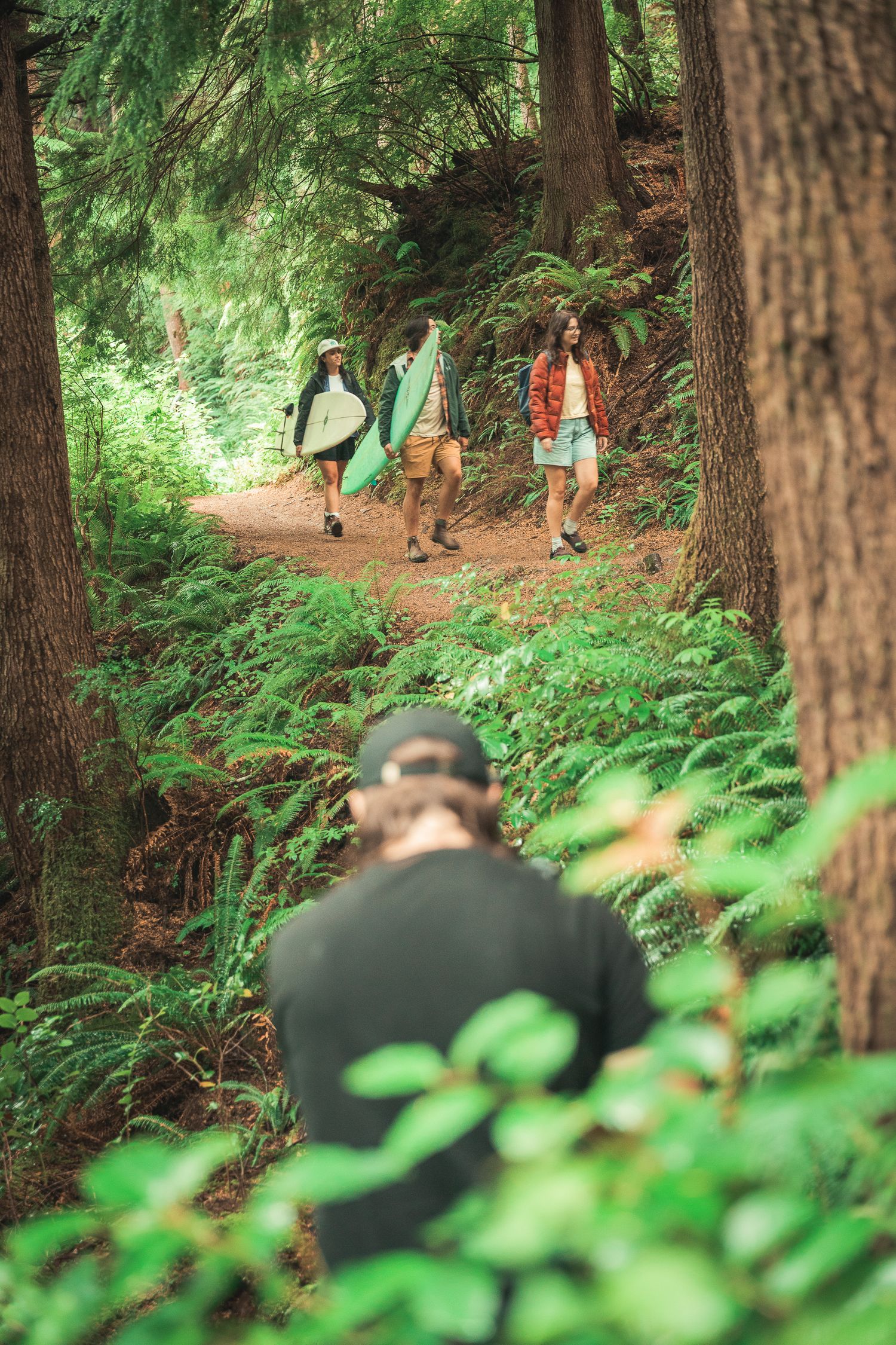Friends carrying surfboards walk a lush forest trail as a photographer captures them from the foreground.