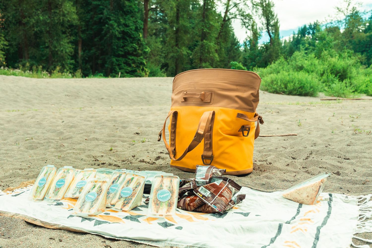 Yellow tote bag and picnic blanket with snacks on a sandy beach surrounded by lush green trees.
