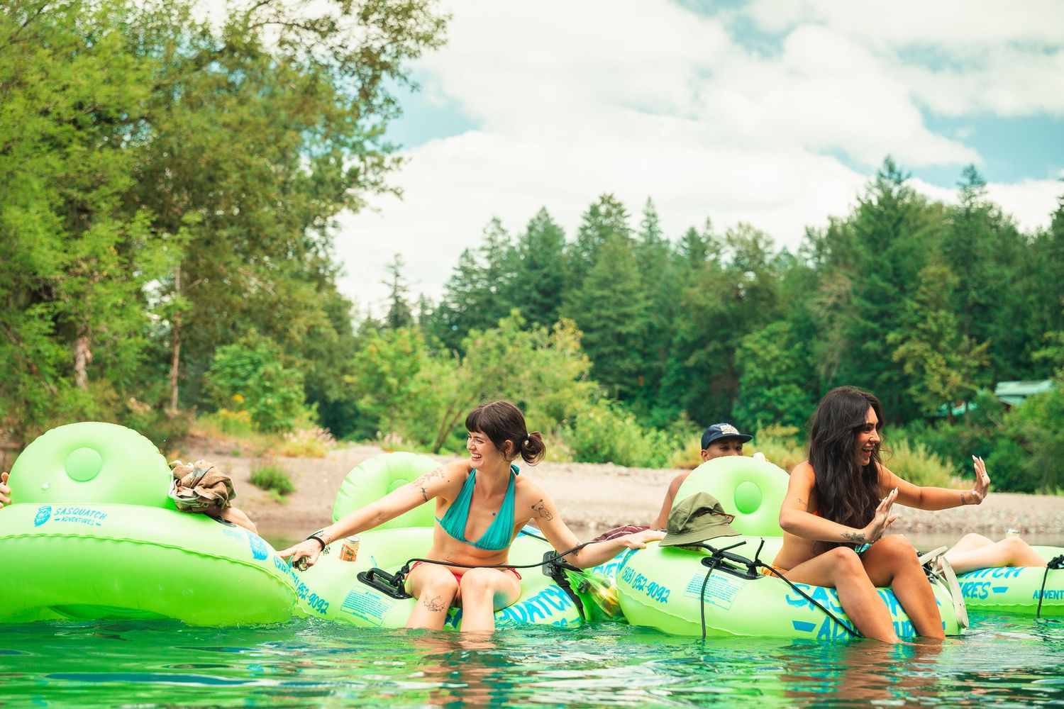 Group of friends enjoying river tubing on bright green inflatable tubes surrounded by lush forest.