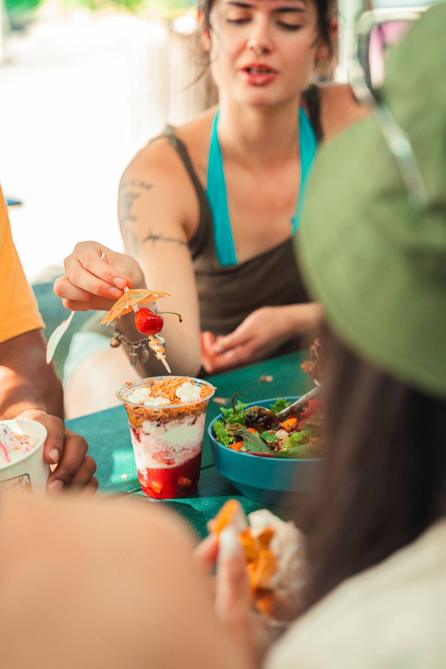 Woman with tattoos serving a strawberry dessert cup at an outdoor food stall with fresh salad nearby.