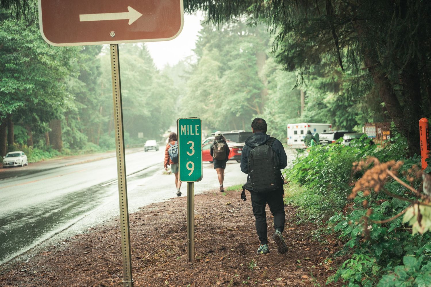 Hikers walking along a rainy forest road past a Mile 39 marker sign surrounded by tall green trees.