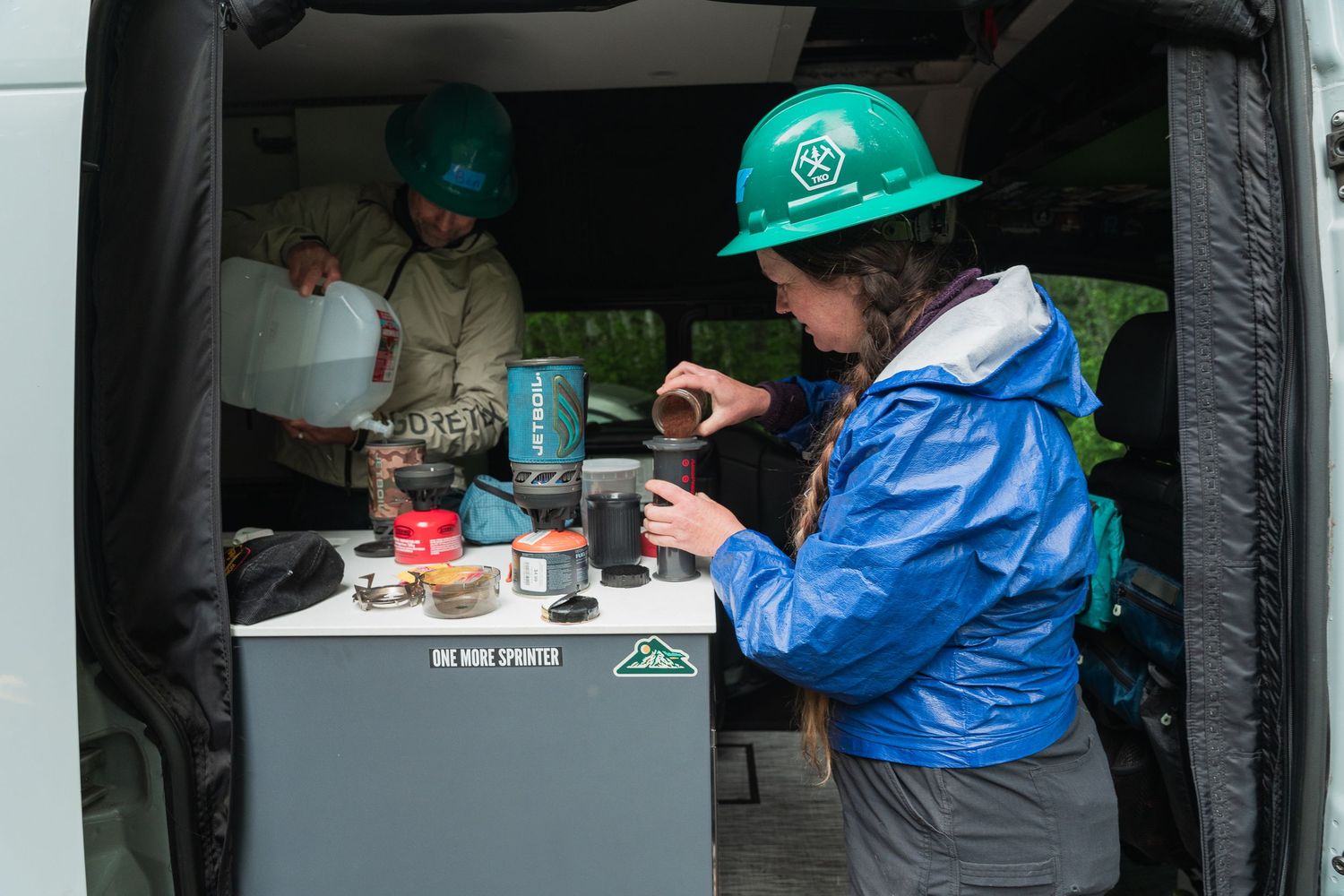 Two people in green hard hats prepare coffee from the back of a van at an outdoor adventure base camp.