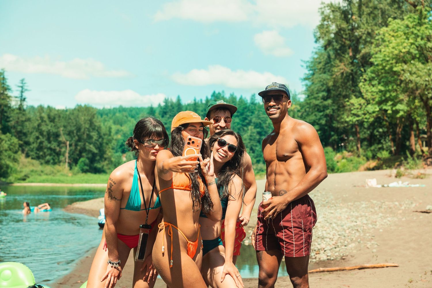 Group of five friends posing for a selfie on a sunny beach by a river, wearing swimwear, smiling and enjoying summer.