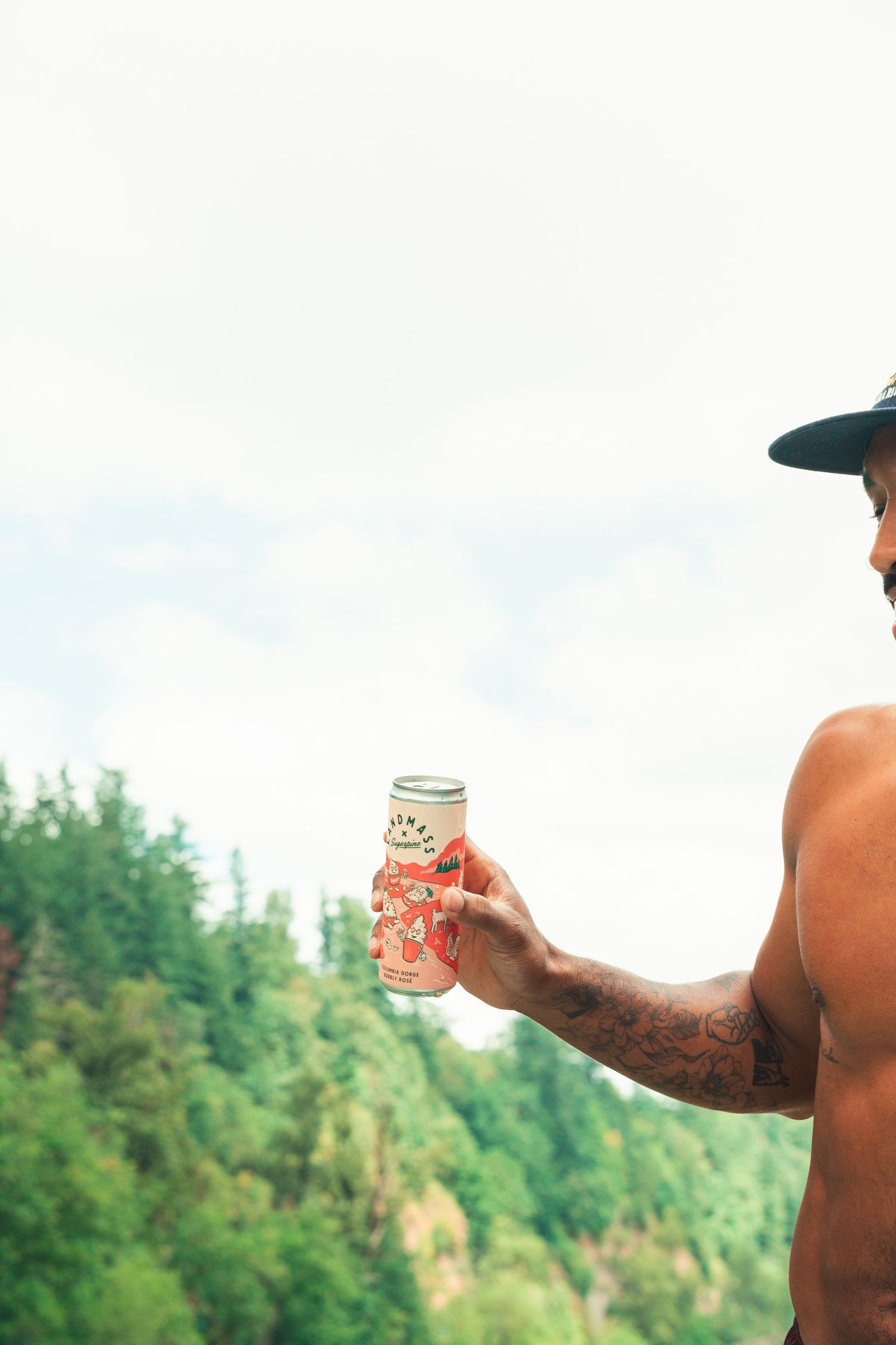 Shirtless tattooed man holding a canned beverage outdoors with lush green forest trees in the background.