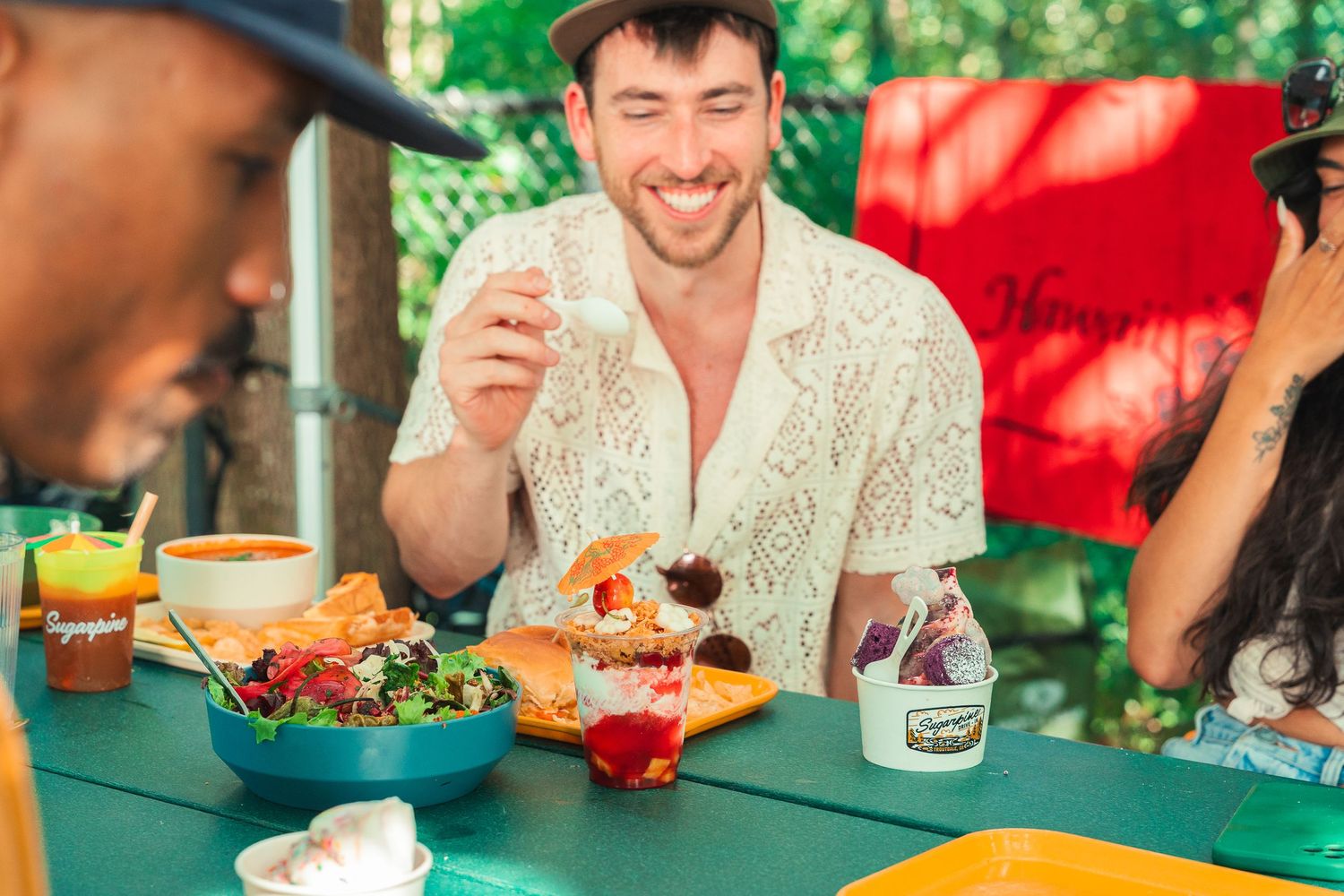 Smiling man enjoying a colorful meal outdoors at a vibrant table with friends in a lush green setting.