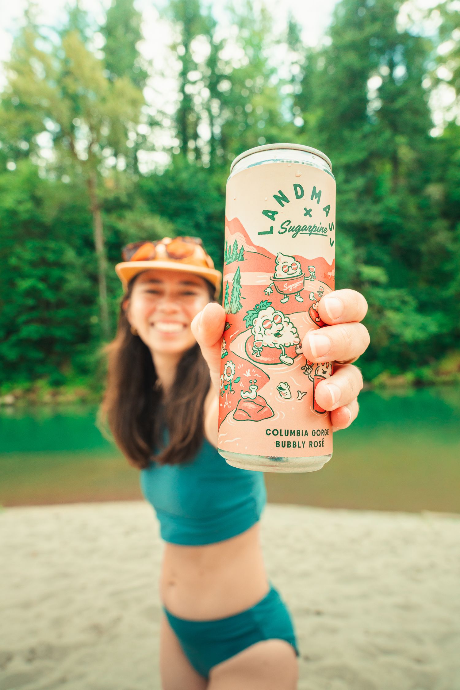 Woman holding a Landm x Sugarpine Columbia Gorge Bubbly Rosé can outdoors near a river.