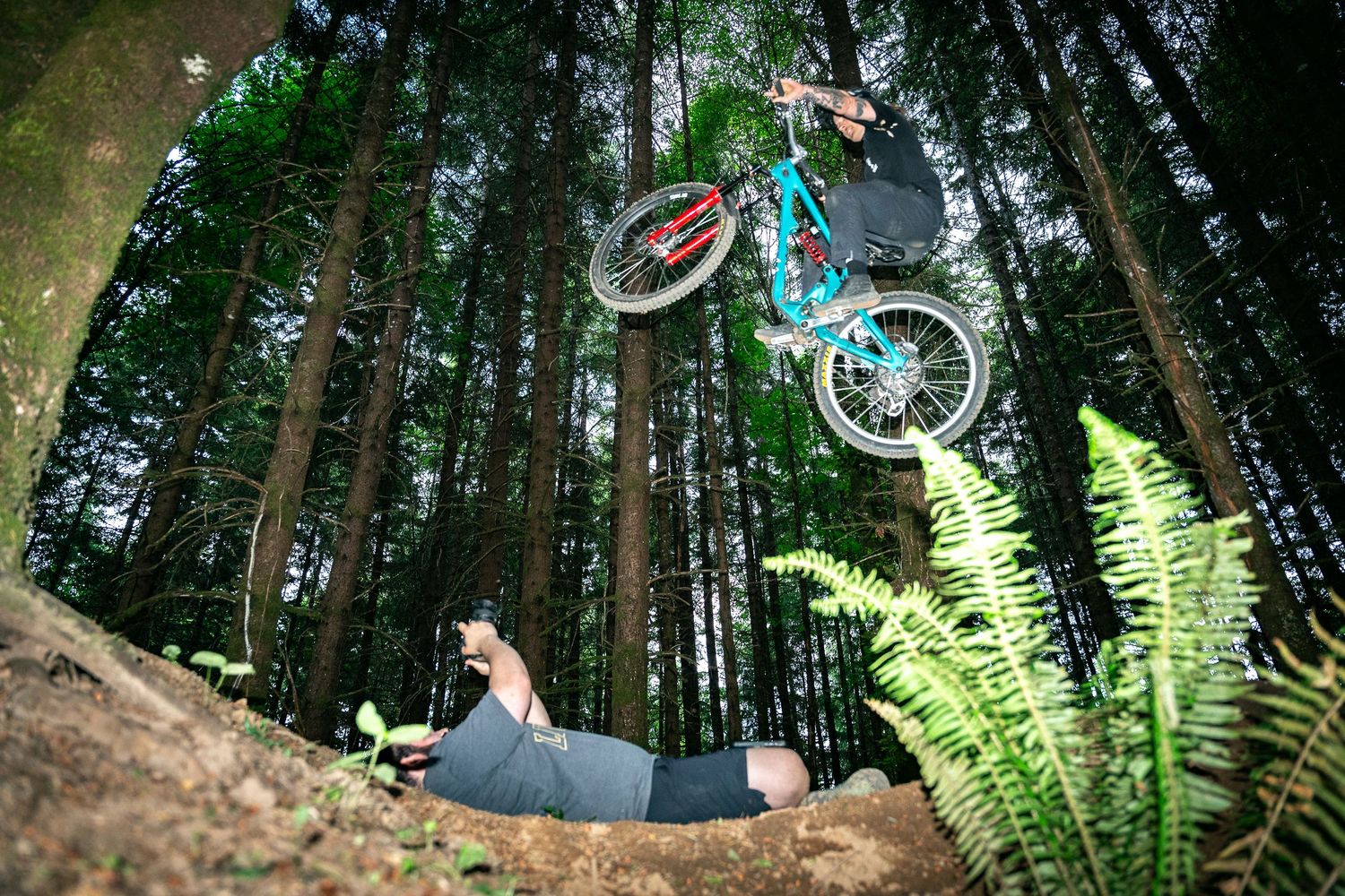 A mountain biker performs a dramatic aerial trick over a person lying on the ground in a dense forest trail.