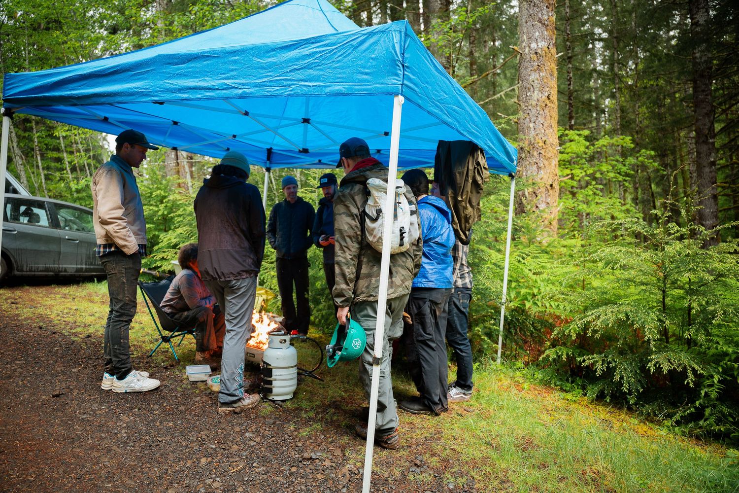 Group of people gathered under a blue tarp canopy in a forest campsite surrounded by tall evergreen trees.