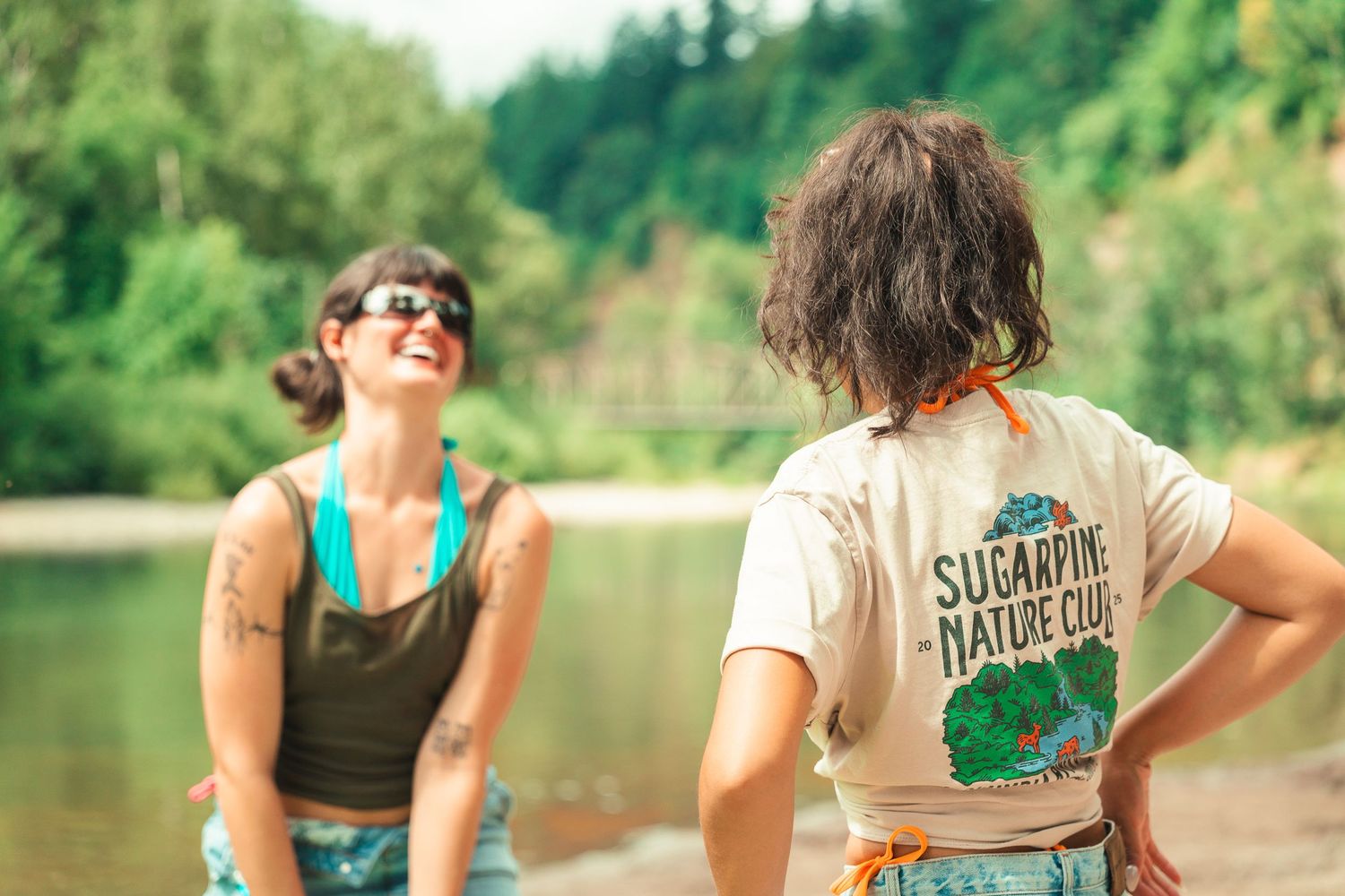 Two women laughing by a river, one wearing a Sugarpine Nature Club t-shirt.