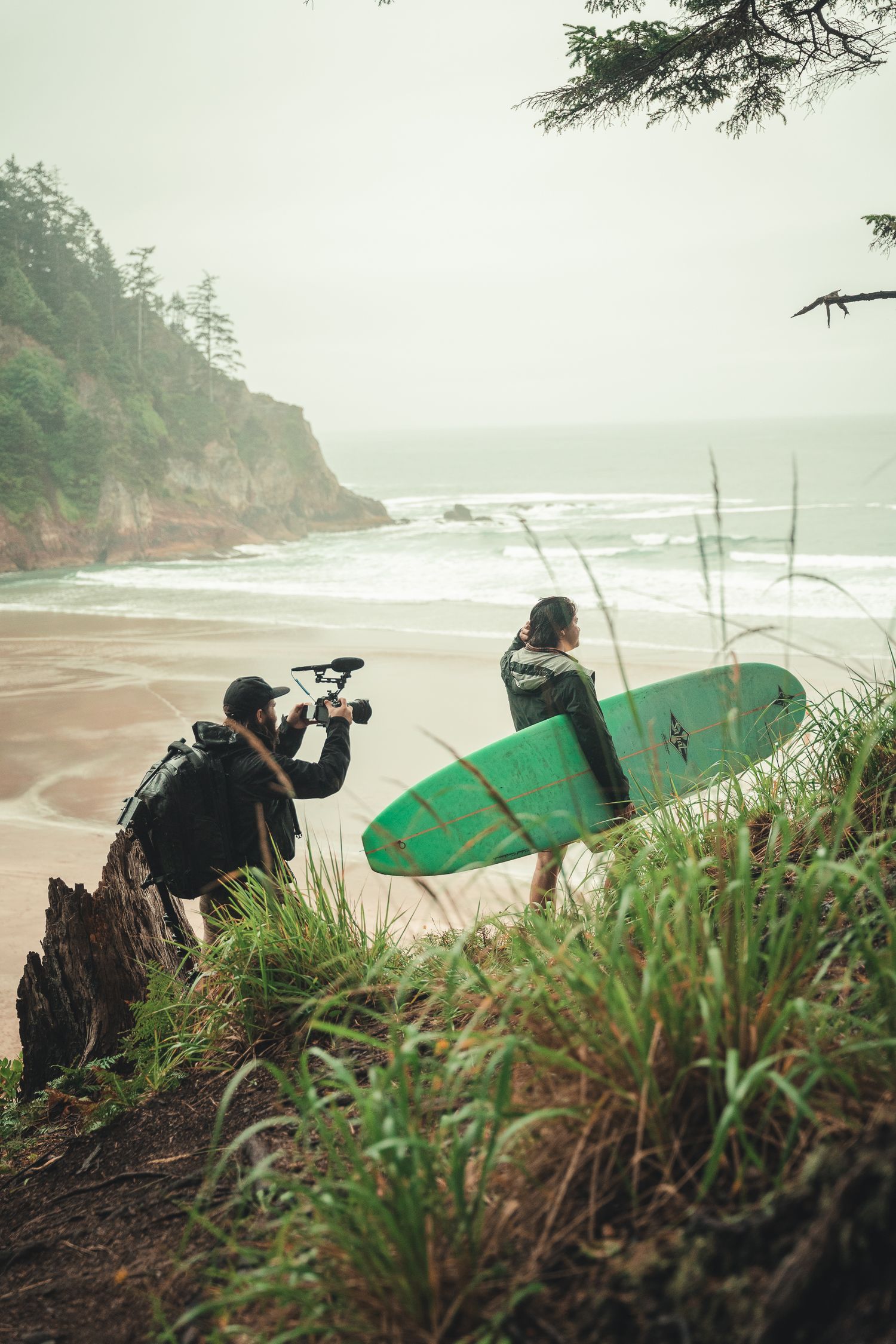 A filmmaker films a surfer carrying a green surfboard on a misty beach with waves and rocky cliffs in the background.