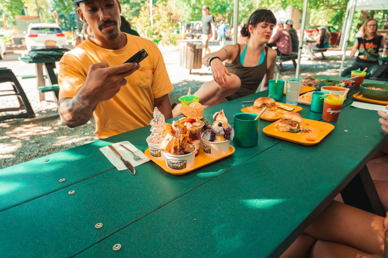 People enjoying colorful ice cream sundaes at a green picnic table outdoors on a sunny day.