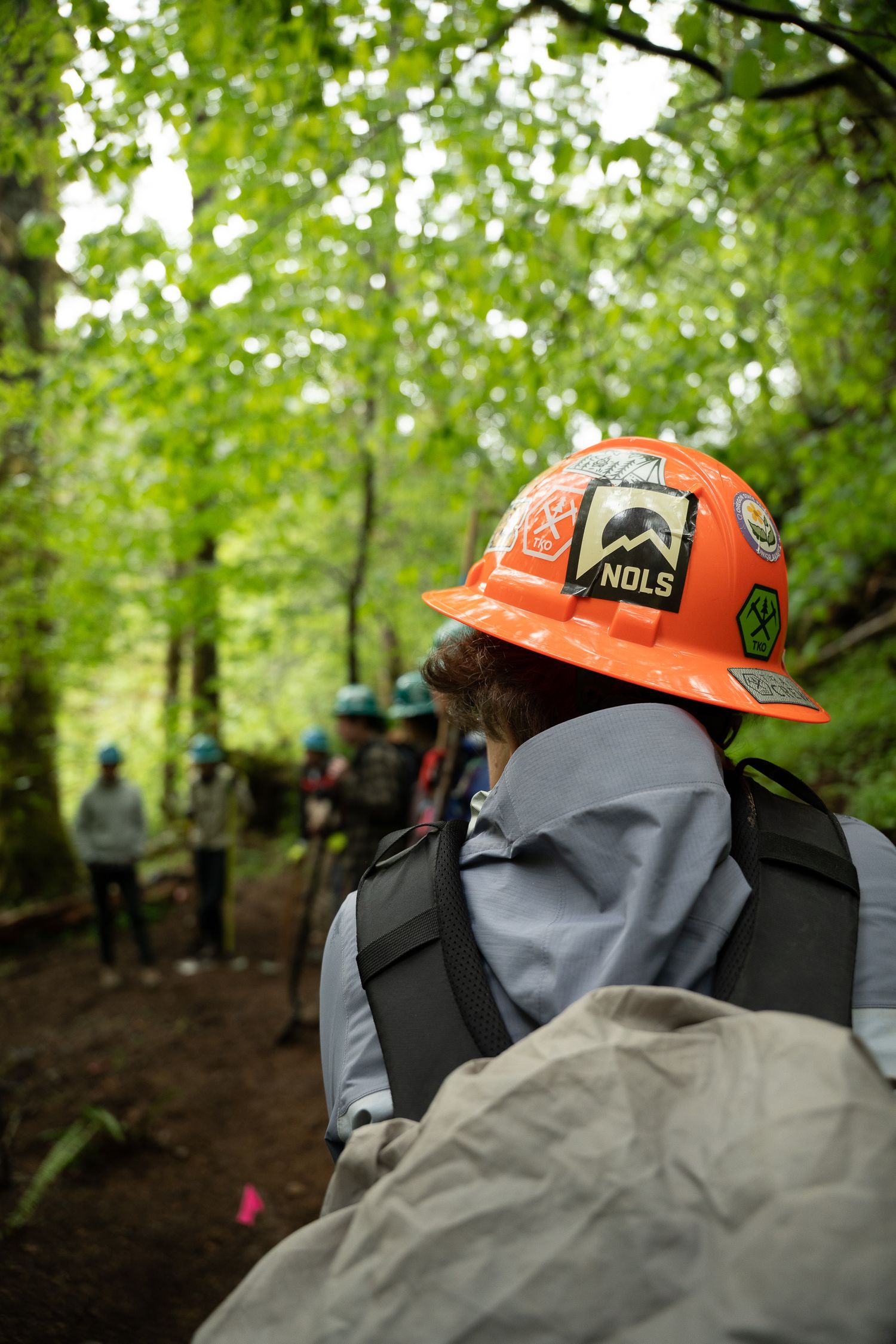 Hiker with orange NOLS hard hat and backpack on a forest trail surrounded by lush green trees.