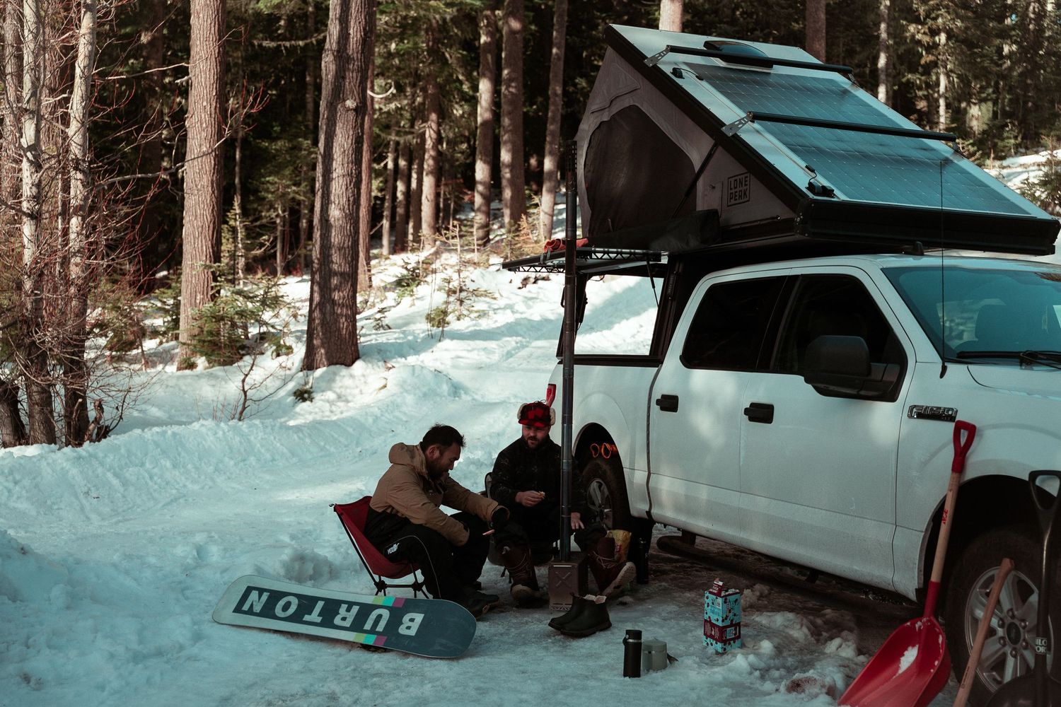 A white SUV is parked in a snowy forest while people set up camp nearby.