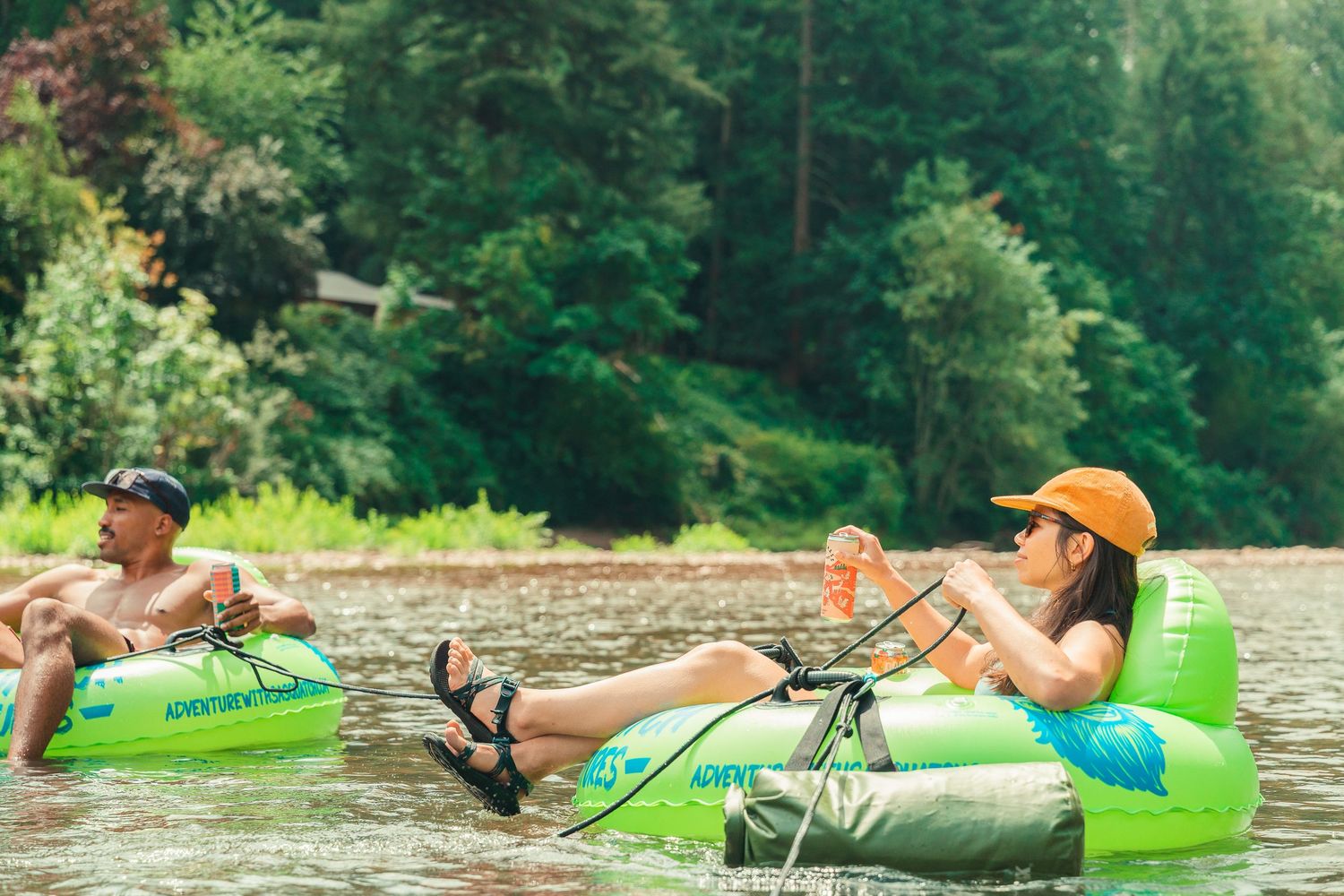 Two people relaxing on green inflatable river tubes floating on a calm river surrounded by lush forest trees.