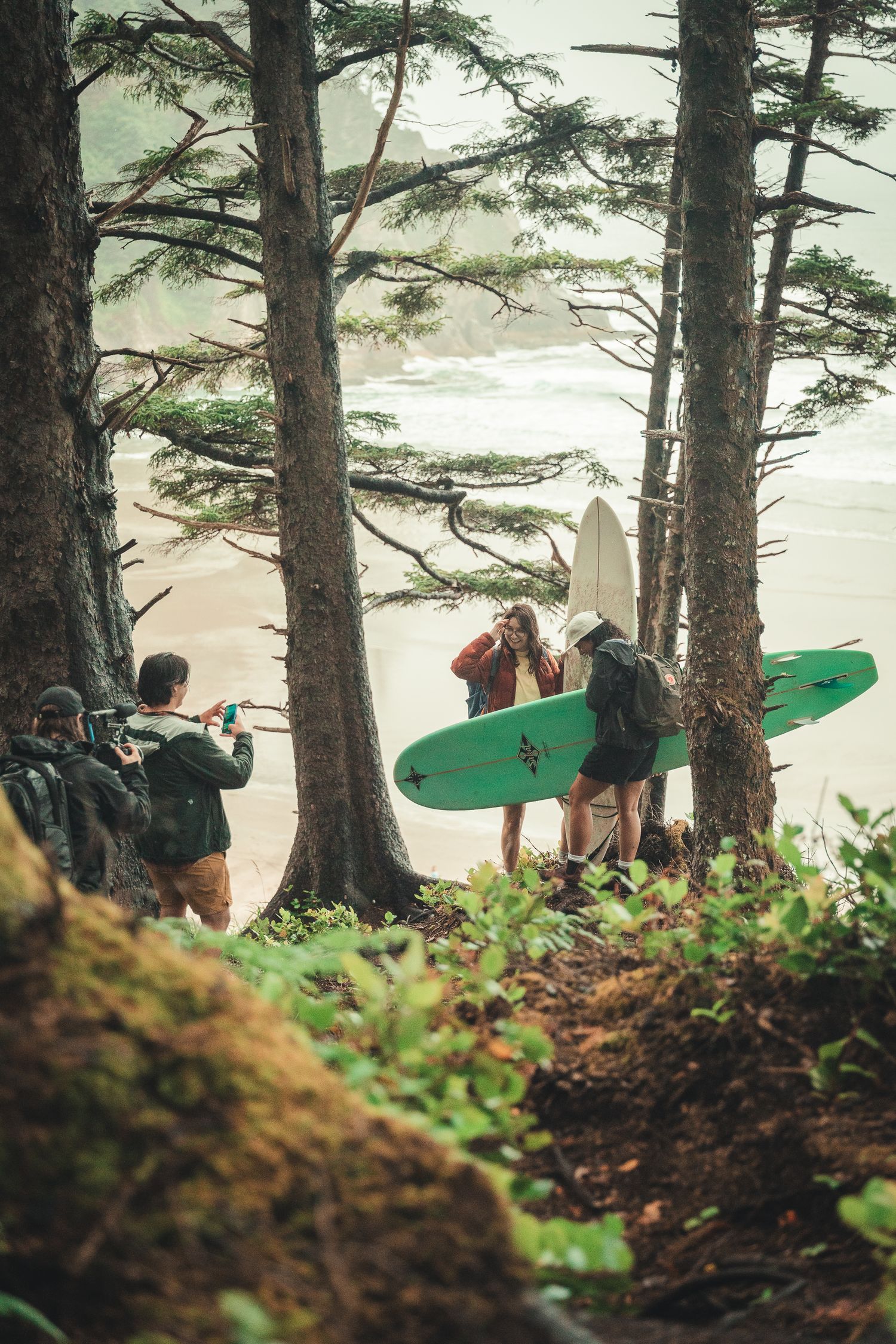 Surfers with boards walk through coastal forest trees toward a misty beach while a photographer captures the scene.