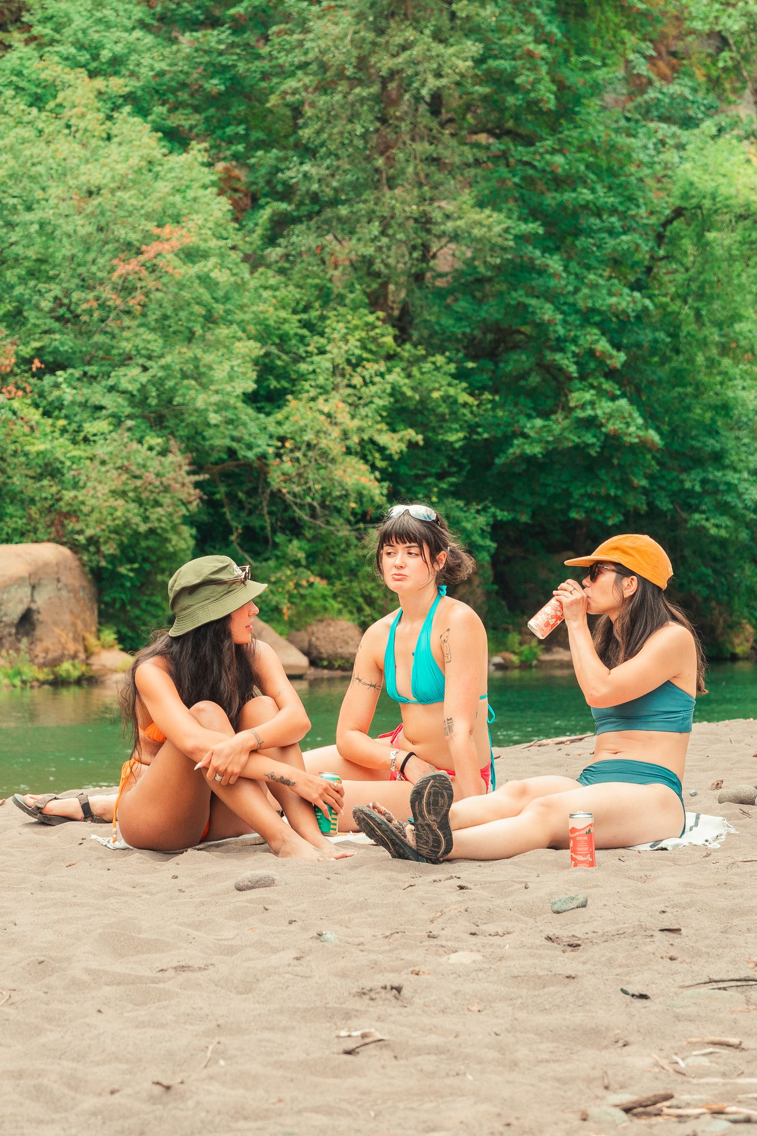 Three women in bikinis and hats sitting on a sandy riverbank surrounded by lush green trees.