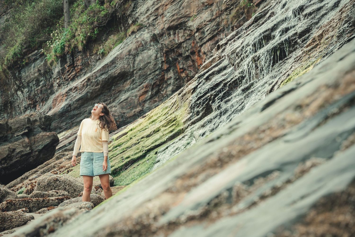 Woman in shorts standing against layered rock cliff face with green moss, gazing upward at scenic coastal formation.