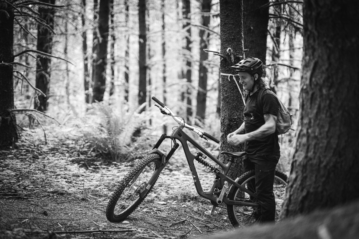 A mountain biker in a helmet stands with their bike on a forest trail in this black and white outdoor photo.