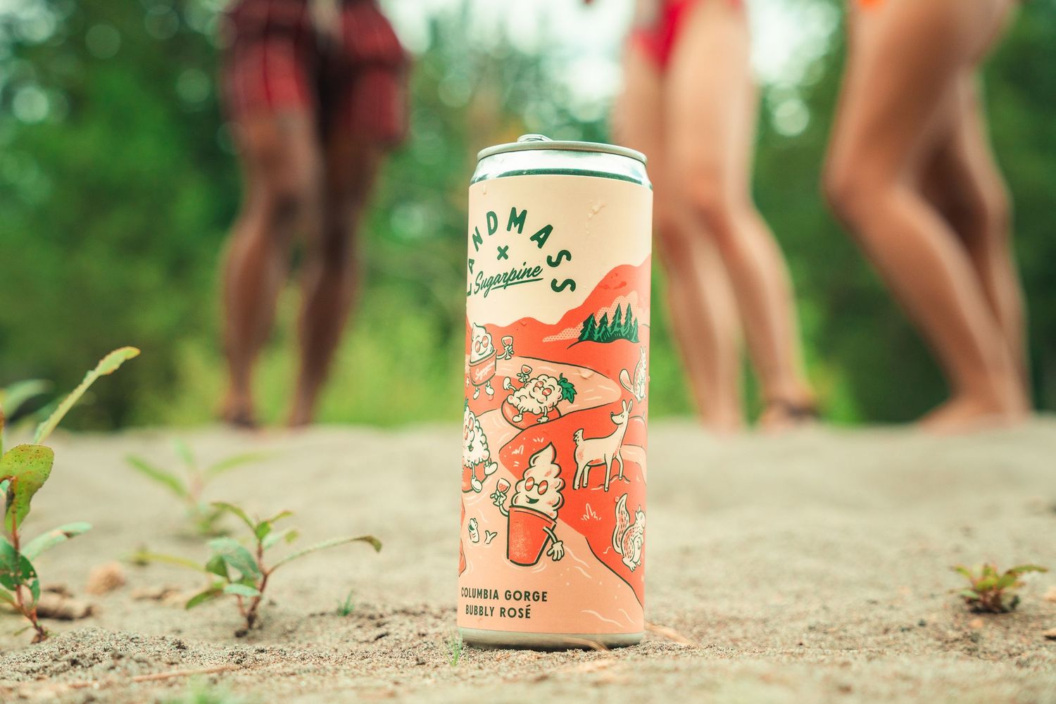 A can of Thomas Champagne rosé sits on sandy ground outdoors with people standing in the background.