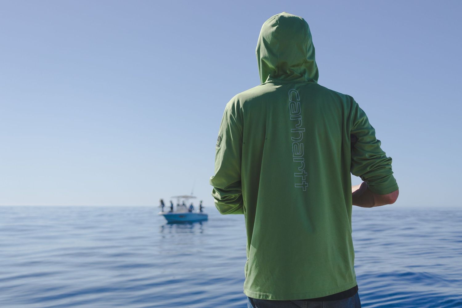 Man in green hooded fishing shirt gazes at a boat on calm blue ocean waters.