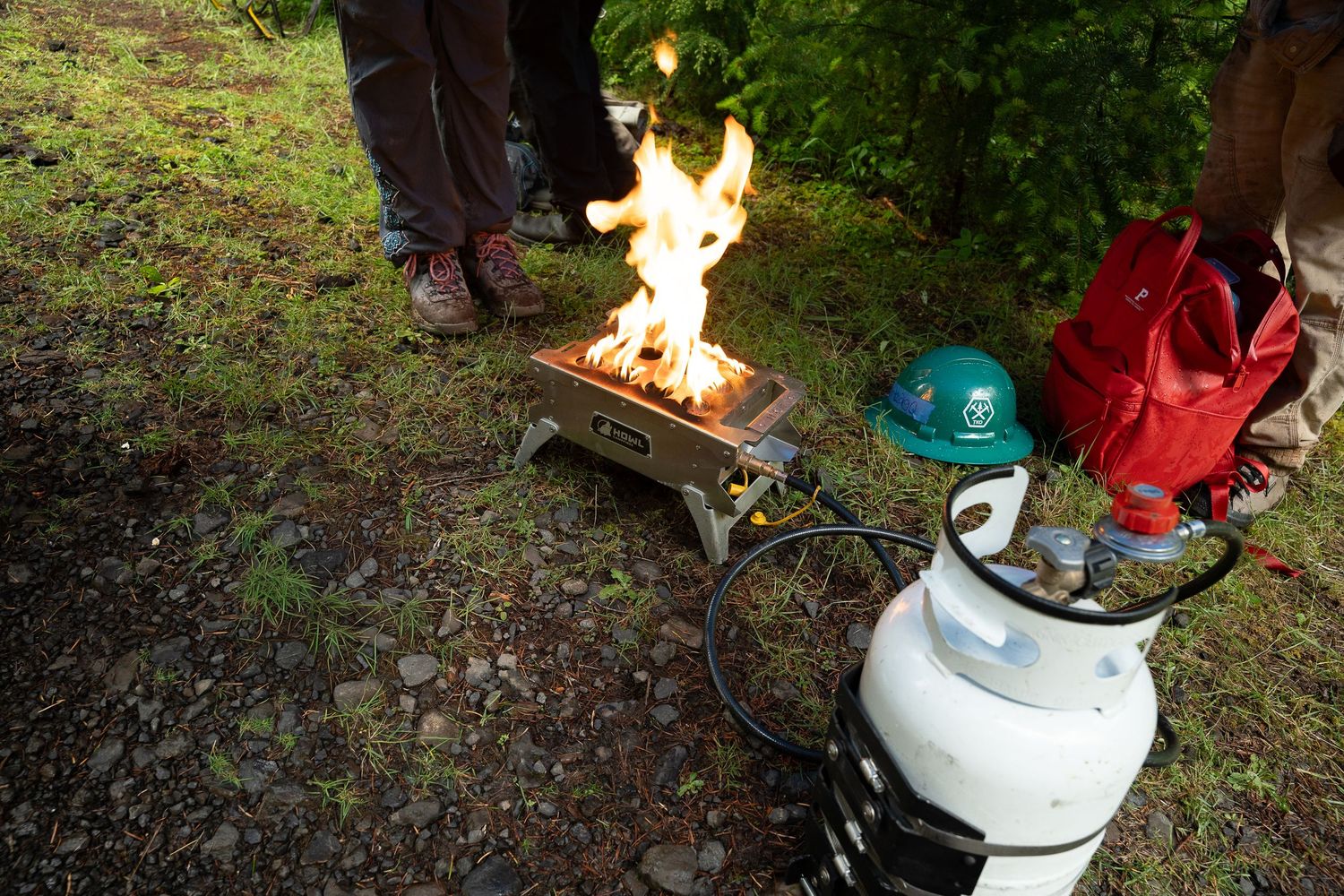 Portable propane camp stove with open flame burning outdoors near green trees and rocky ground.