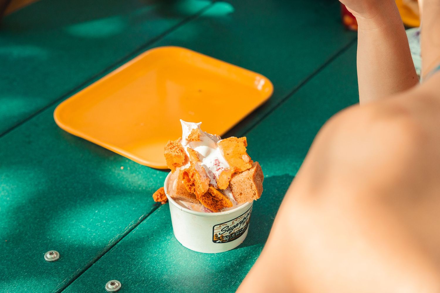 A cup of orange ice cream with toppings on a green picnic table beside a yellow tray in bright sunlight.