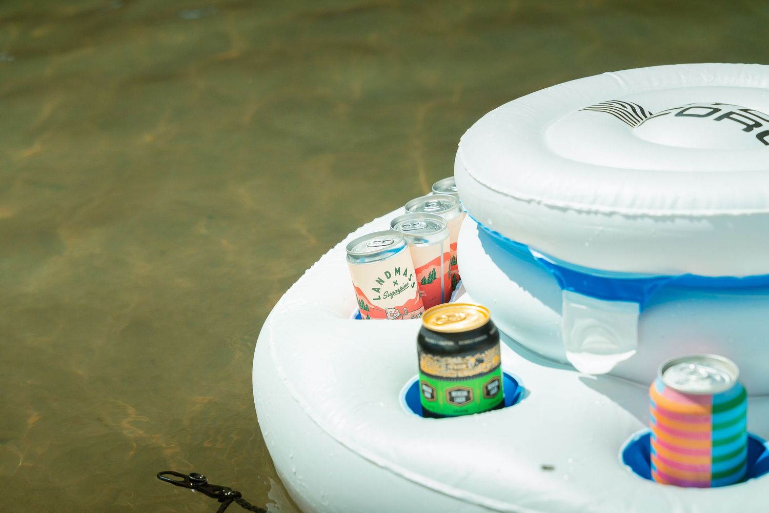 Inflatable pool float bar with colorful canned drinks and shot glasses floating on water.