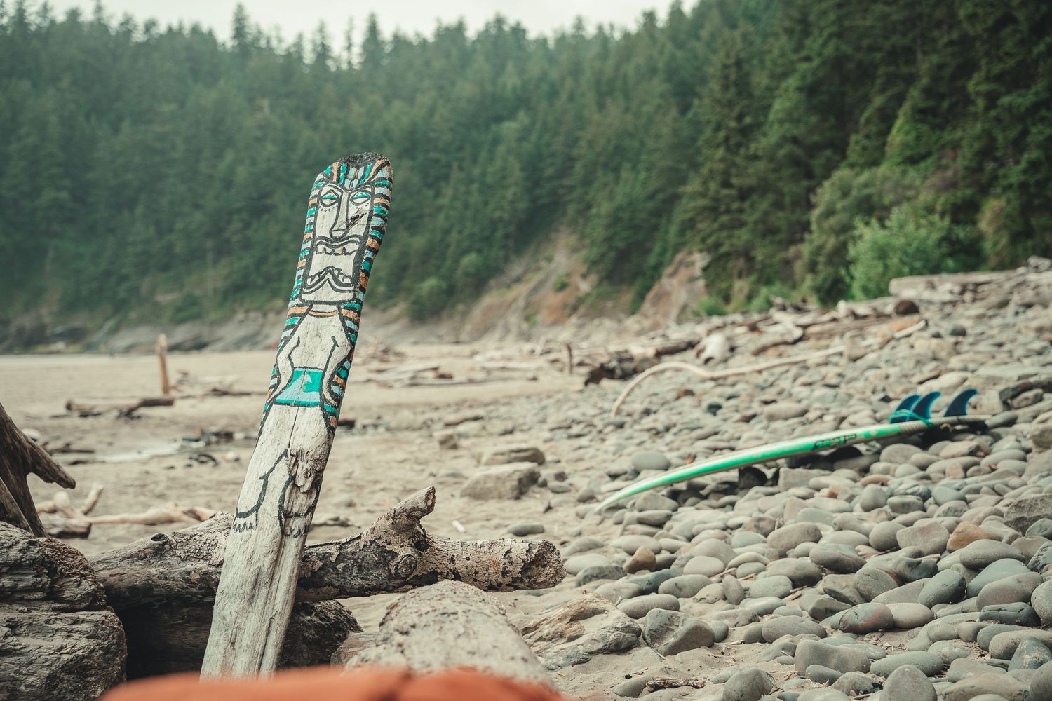 Decorated driftwood totem on a rocky Pacific Northwest beach with surfboards and dense forest backdrop.