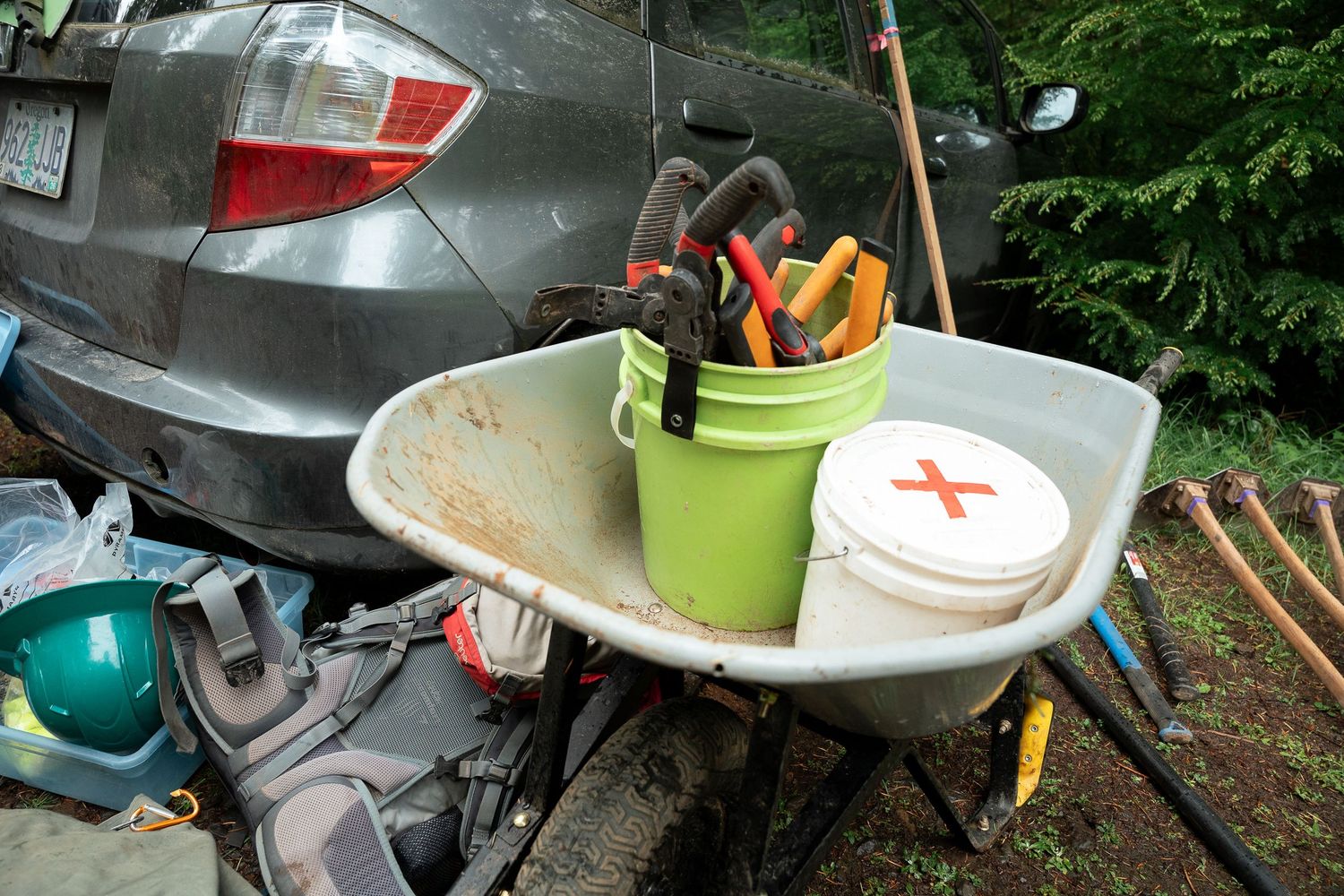 Wheelbarrow holding green bucket with tools and white bucket near gray car outdoors.