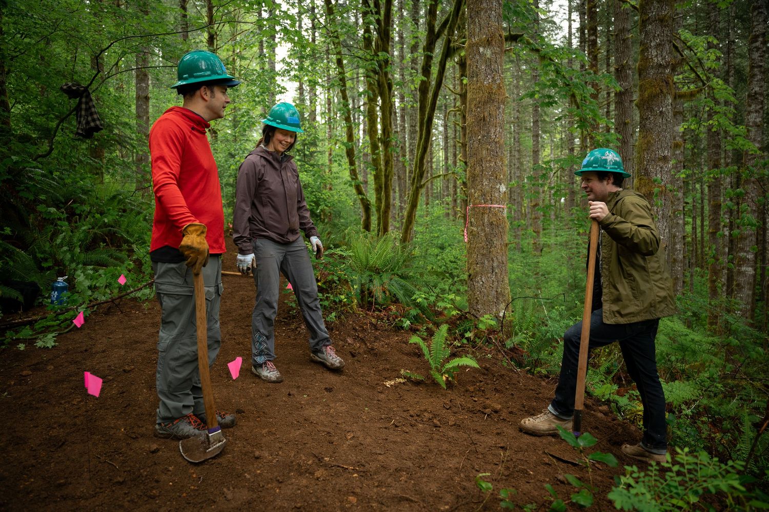 Three workers in green hard hats and gloves use shovels to build a forest trail surrounded by lush green trees.