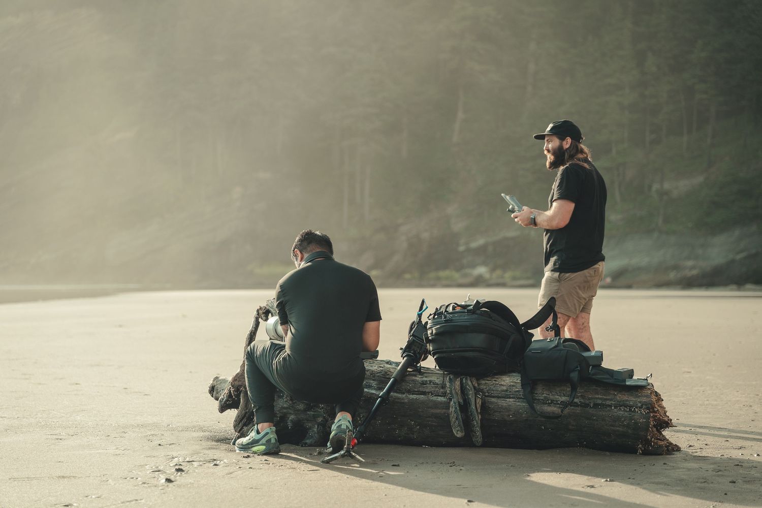 Two hikers with backpacks resting on a log on a sandy beach surrounded by misty forested mountains.