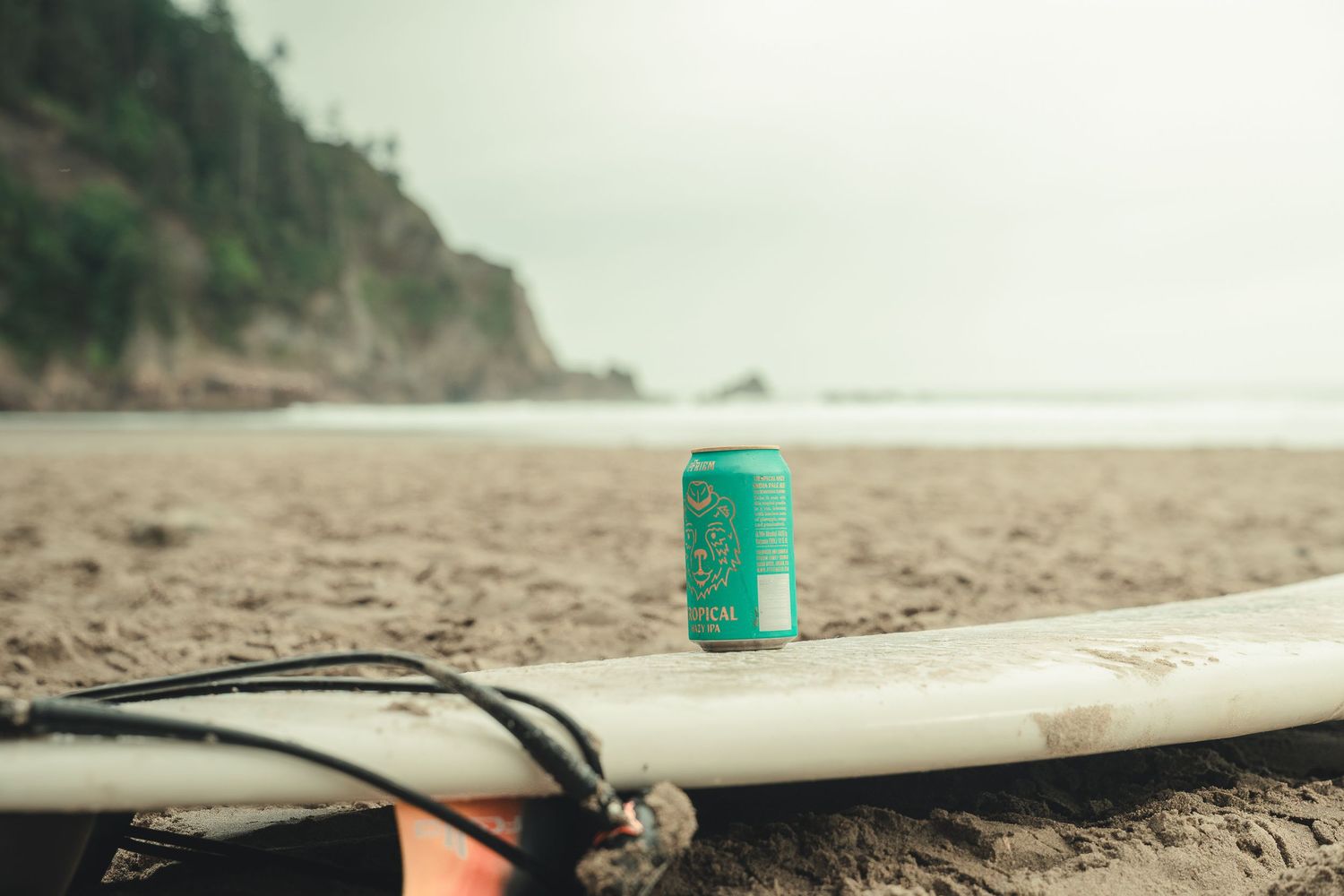 A green beer can sits on a white surfboard on a sandy beach with a rocky cliff in the background.