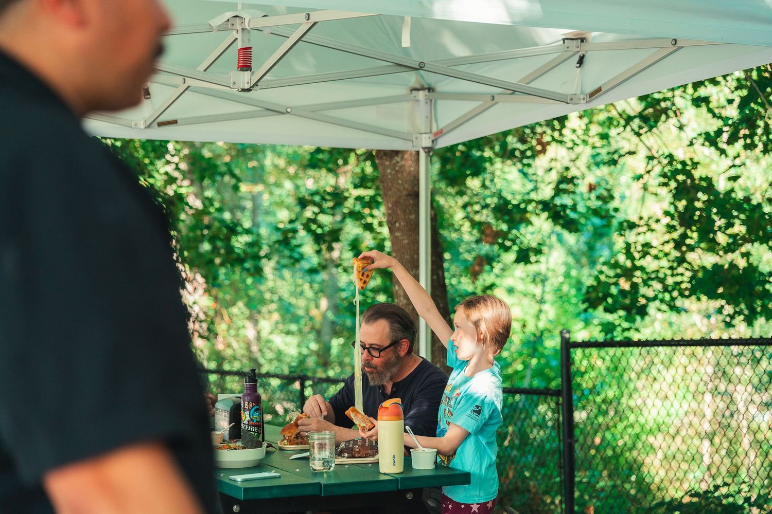 Man and child dining outdoors under a white umbrella canopy surrounded by lush green trees.