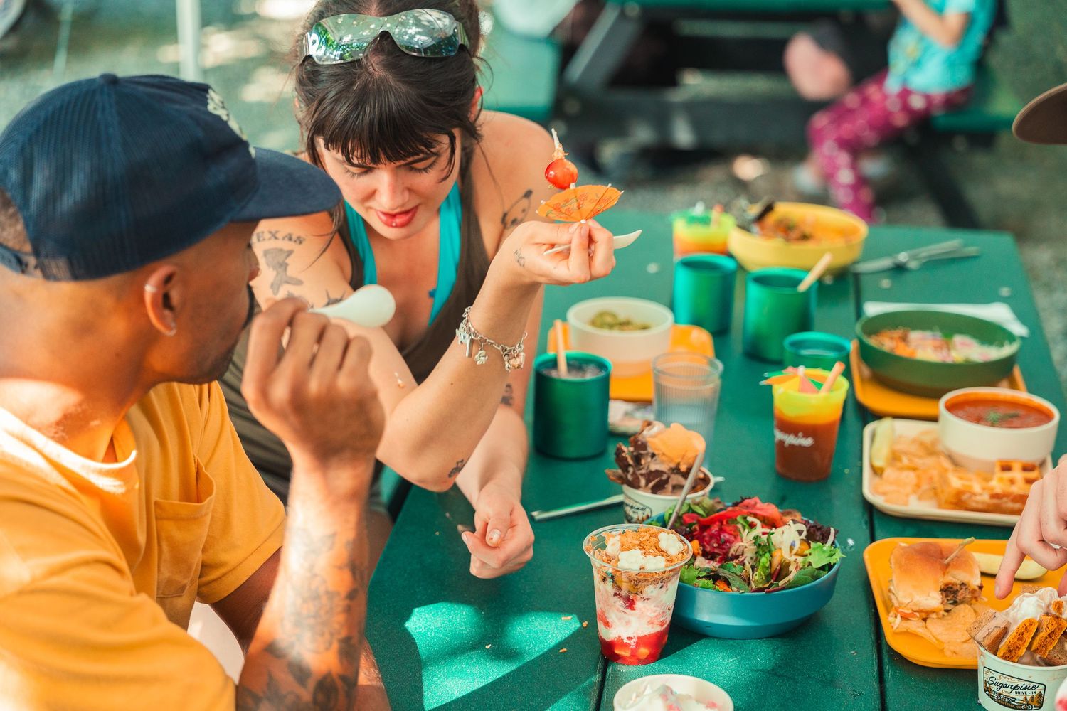 Friends enjoying a colorful outdoor meal together at a green picnic table with various dishes and drinks.