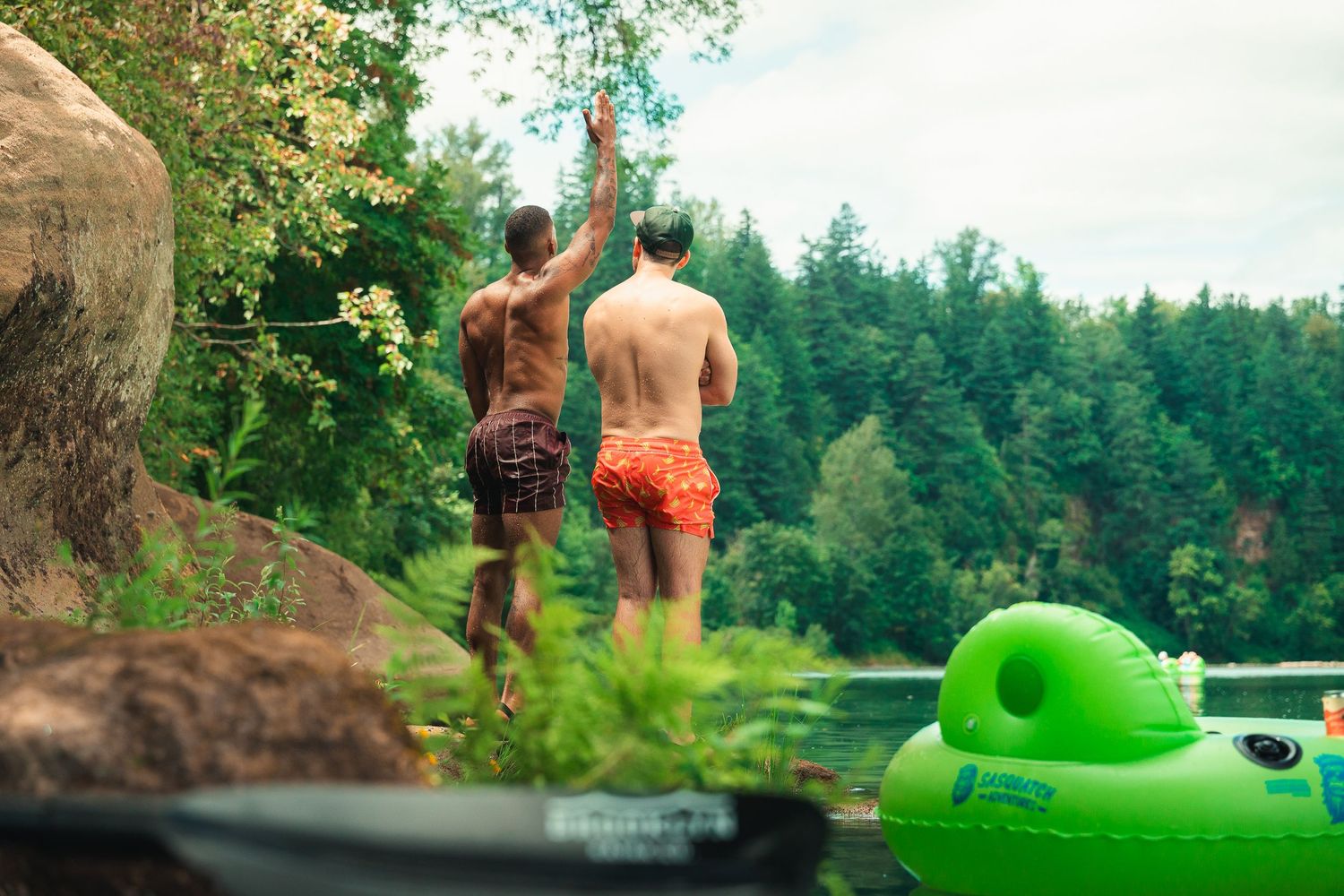 Two men in swim trunks high-fiving near a lake with a green inflatable float and lush forest backdrop.
