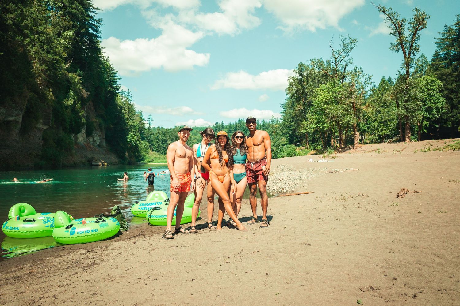 Group of friends posing on sandy riverbank with green inflatable tubes, surrounded by lush forest trees on a sunny day.
