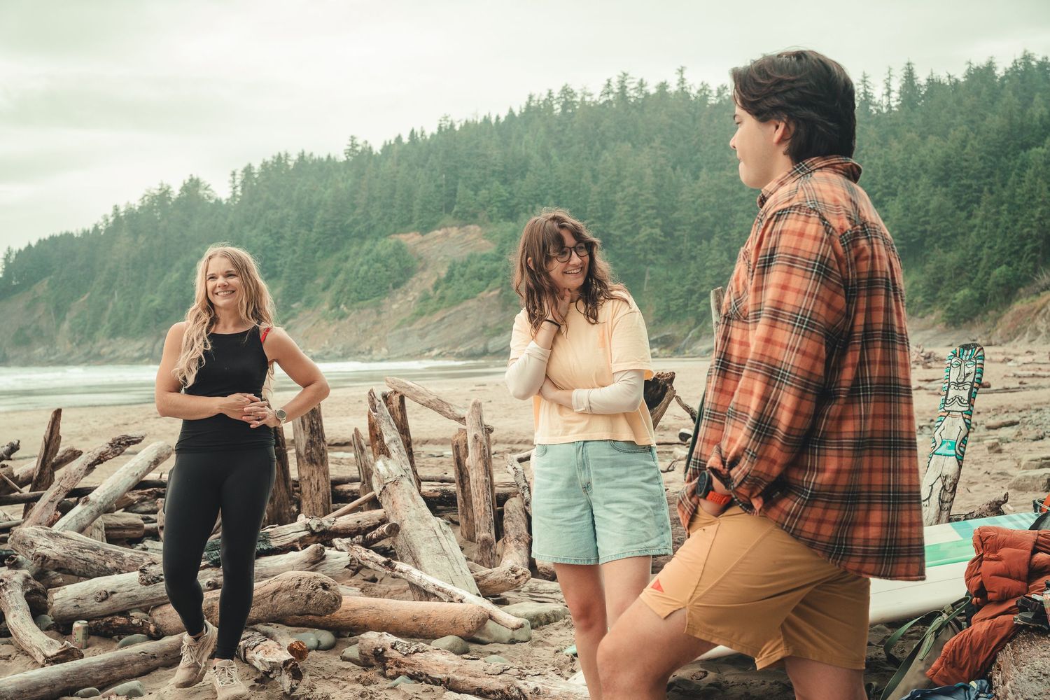 Three people socializing on a driftwood-covered Pacific Northwest beach with lush green forest hills in the background.