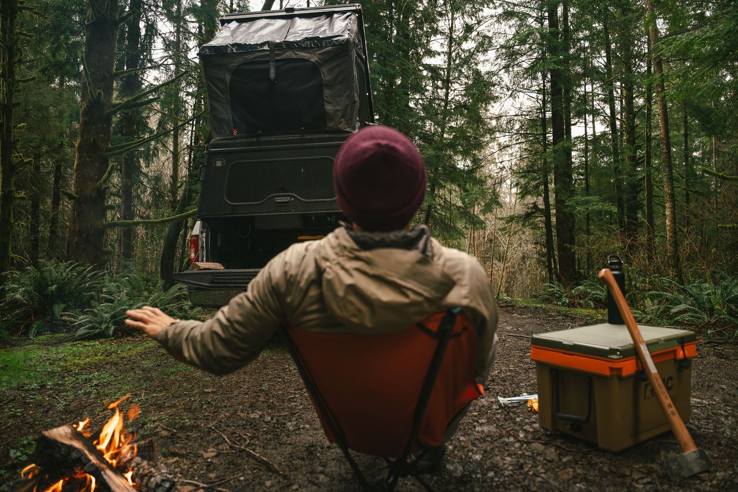 Camper relaxing by campfire near vehicle with rooftop tent in dense forest.