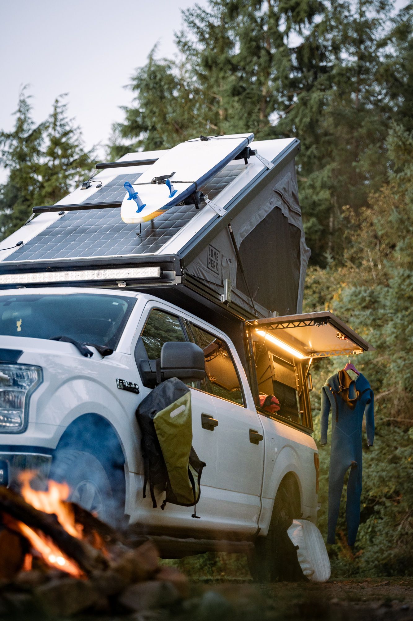 White truck with rooftop camping setup and solar panels parked in a wooded area with campfire glow.