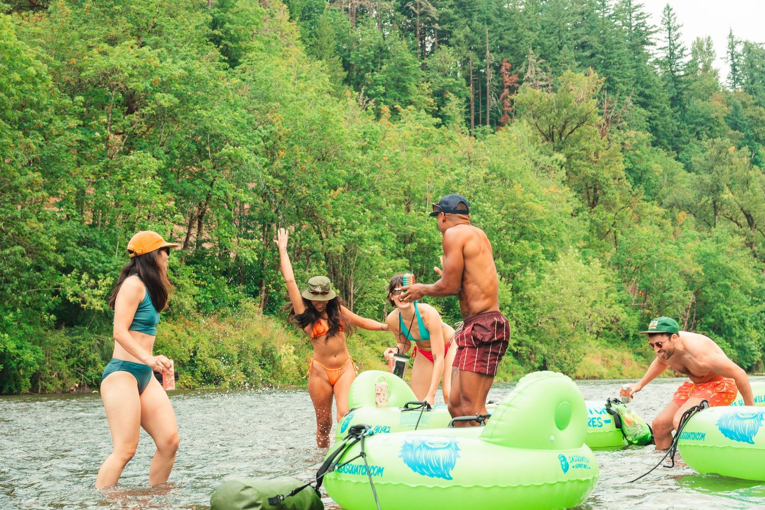 Group of friends in swimwear enjoying a river day with inflatable tubes surrounded by lush green forest.