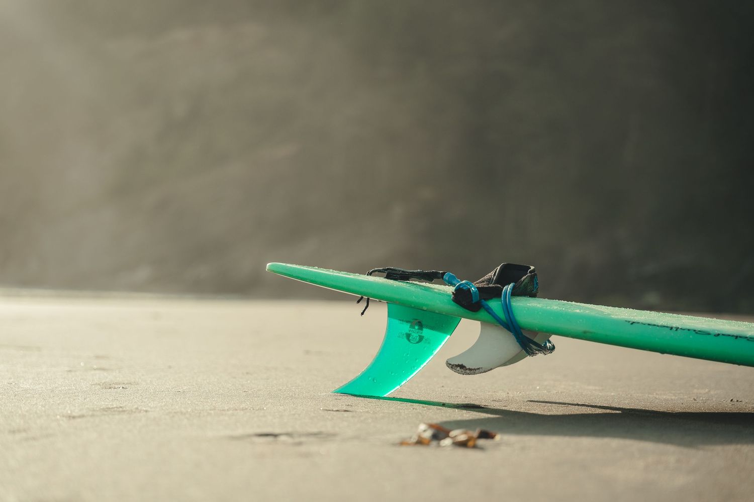 A teal surfboard with fins resting on a sandy beach in soft, hazy natural light.