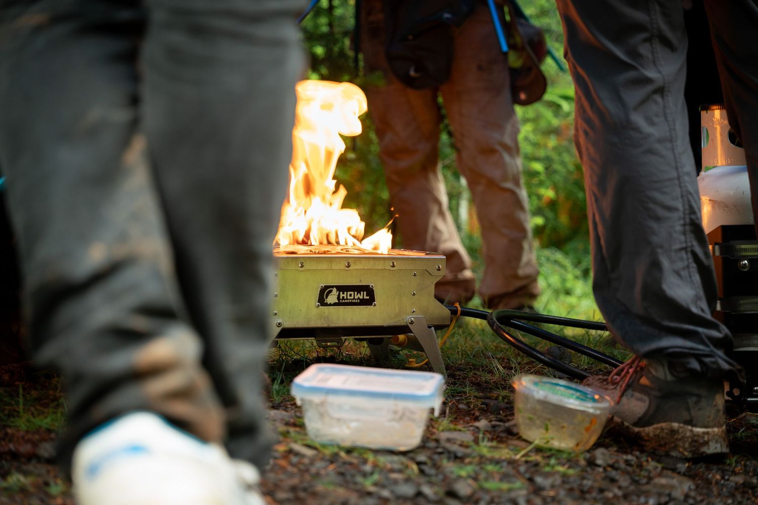 Group camping with a portable stove producing flames outdoors, surrounded by food containers on rocky ground.