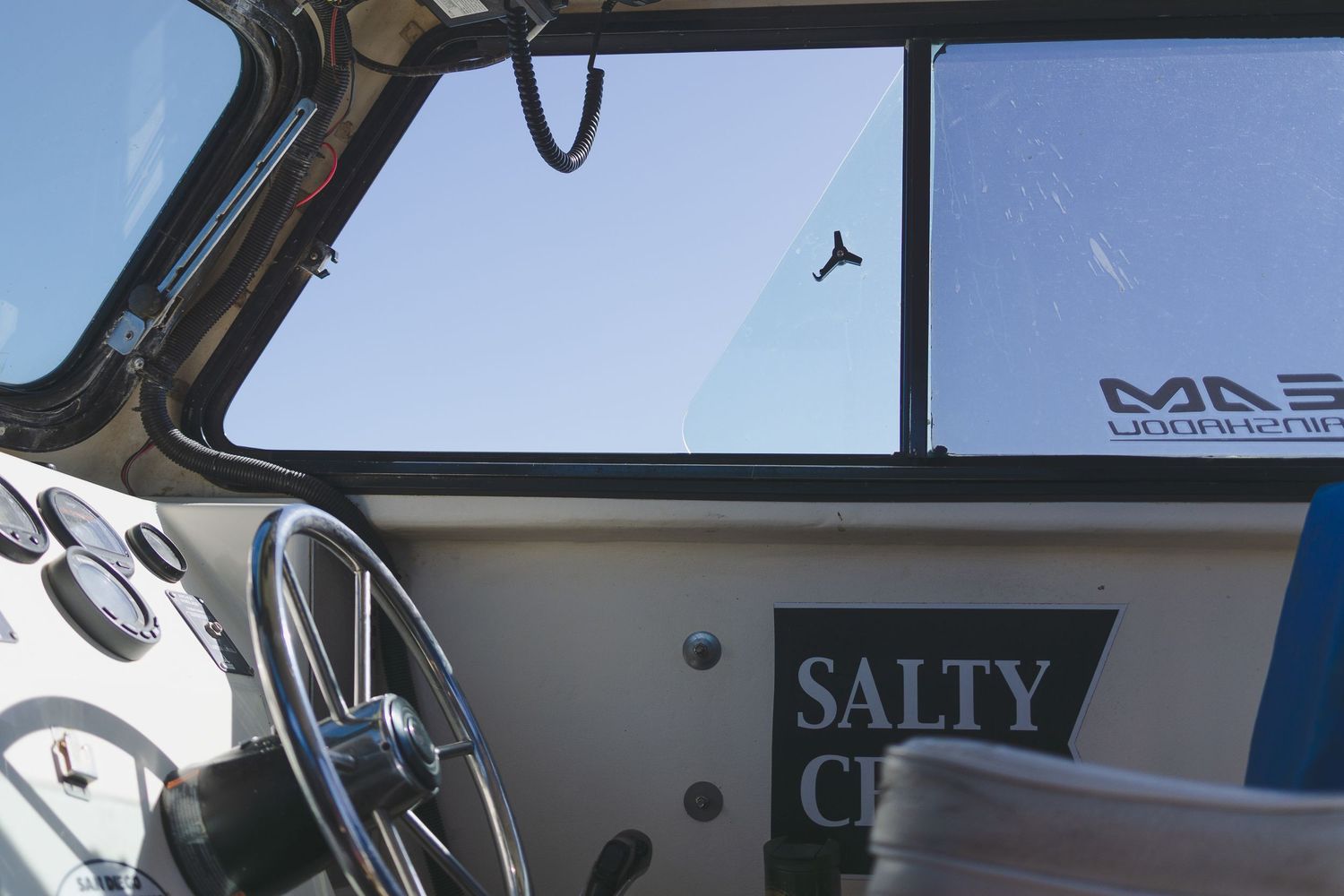 Interior view of a boat cockpit with steering wheel, Salty C sticker, and clear windshield under blue sky.