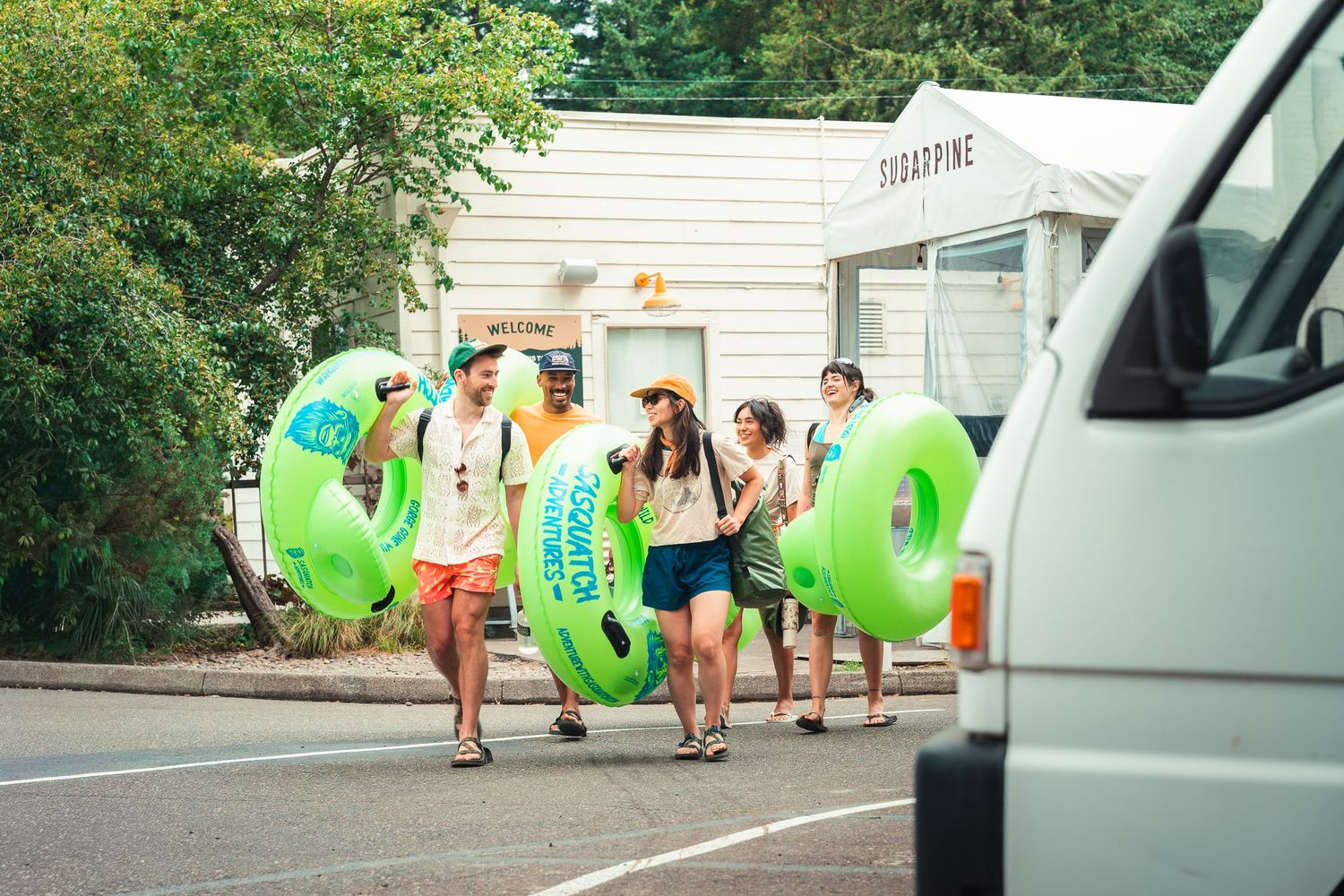 Group of friends walking outdoors carrying large green inflatable pool floats near a white van on a sunny day.